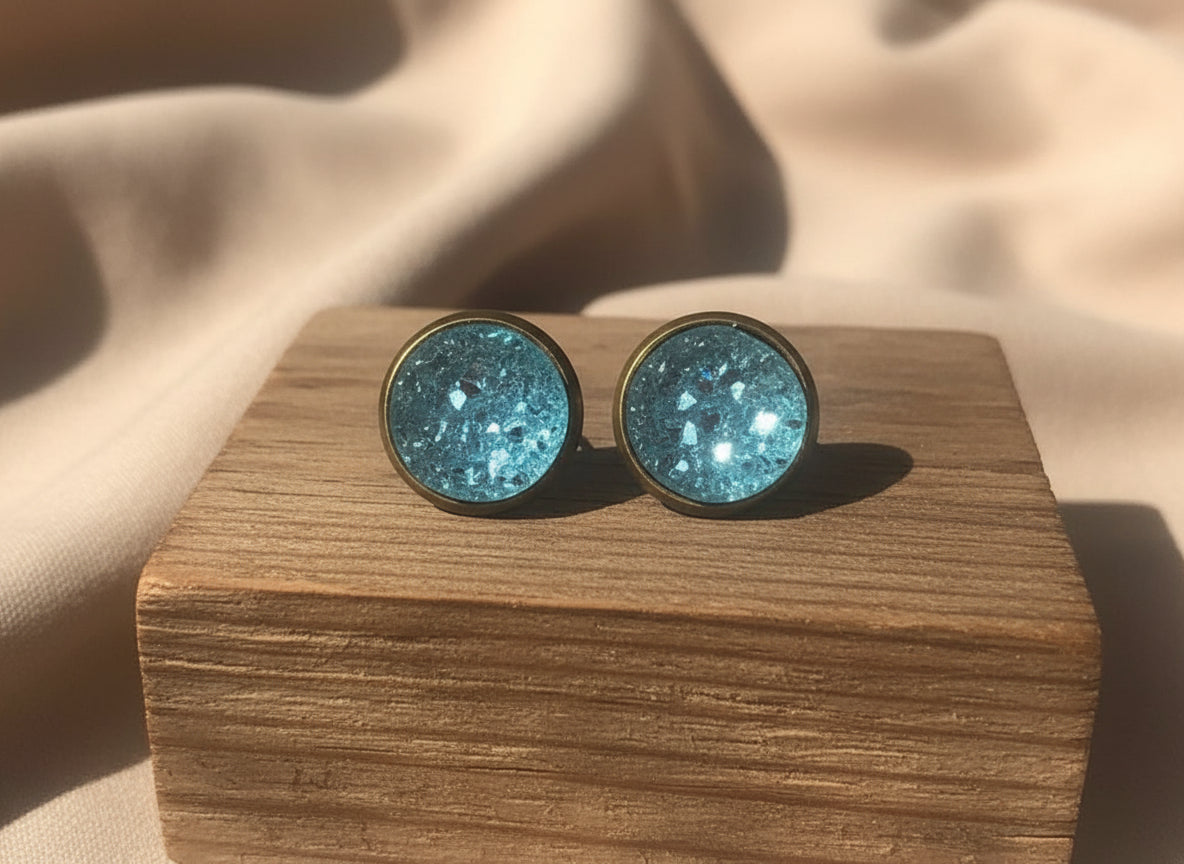 Drift Glass druzy stud earrings in soft aqua blue with crystalline shimmer, set in metal studs on a wooden block